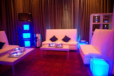 A section of the St. Regis Astor ballroom transformed into a Smartwater lounge for the evening, complete with white couches and curtains, a flat-screen TV with a branding video on a loop, and glowing blue plastic cube tables.