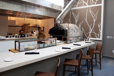 Near the restaurant entrance, a four-seat dining counter faces an open kitchen and wood-burning oven.