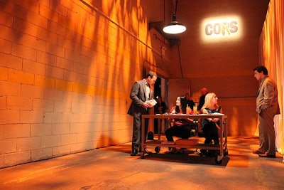 Rather than the venue's main entrance, the event used the loading dock to check in guests, setting the tone for the evening with the space's bare walls and concrete floors. Decor in this area included a plain, stainless steel table, a single hanging light fixture, and a 'cops' sign defaced to read 'cons.'