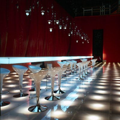 The cocktail area featured a long, gauzy red curtain, six floor-to-ceiling framed mirrors bearing the Vuitton crest, white leather ottomans, and a long white-lit Lucite bar. (Photo courtesy of J. Ben Bourgeois Productions)