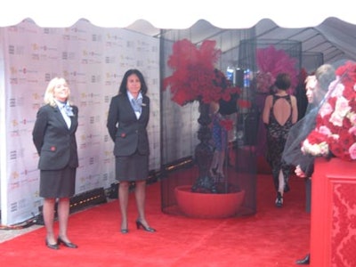 Volunteers stood near the entrance to the event surrounded by large design pieces including black wrought iron pedestals with red flowers and feathers atop and red square carved pedestals with pink and red flowers on top. Fish nets accented both.