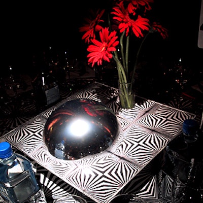 The unusual table centerpieces consisted of silver steel globes and small acrylic vases filled with red gerbera daisies held together by a sheet of clear plastic with the same design of the tablecloth.