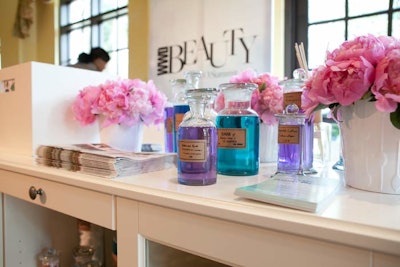 The apothecary-inspired registration desk had different-sized jars filled with colored liquids, cotton balls, and Q-tips. Pink peonies in white vases, assorted bright-colored towels, diffusers, and WWD magazine issues also adorned the desk.