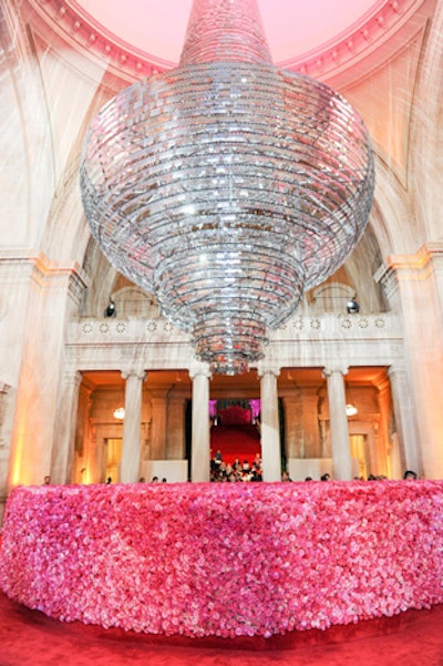 In the center of the museum's Great Hall, a 40- by 24-foot chandelier made by London-based artist and designer Simon Kenny was constructed from 14,000 aluminum 'razor blades,' each 4- by 5-inches in size. The installation hung above the information desk, which was covered in three different shades of pink roses set five feet high all around. Two months in the making and having arrived from London via boat in about 25 pieces that were then constructed on site, the metallic nature of the chandelier allowed it to shine, lit by beams of light from the balcony. A red carpet and cherry-blossom trees flanked the chandelier on all four corners. Guests then proceeded up the main staircase directly behind.