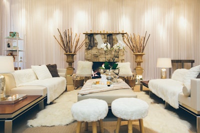 The plush lounge area is filled with couches draped in fluffy, furry white blankets. A prop fireplace adorned with stockings anchors the space, and gauzy white drapes form a wintry perimeter.