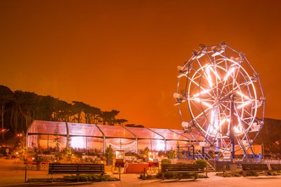 Clear tent illuminated in golden amber glow at outdoor celebration, complete with ferris wheel