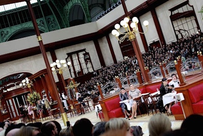 After their two catwalk turns each, models took predesignated seats at a number of padded deep red booths positioned at the center of the runway. They were greeted by waiters in black waistcoats, long aprons, and menus, and the tables were decorated with clusters of globe lamps, white tablecloths, and table réservée signs. Food was then served.
