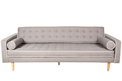 Blueprint Studios' Lolland sofa features pale gray fabric outlined in white piping, along with tufted cushions and roll pillows. The seating is available nationwide; pricing is available on request.