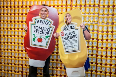 Guests posed with oversize bottles of ketchup and mustard in front of a wall comprising hundreds of bottles of yellow mustard.