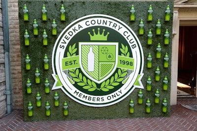 Bottles and a crest emblazoned with 'members only' decorated a press wall. Turf covered the backdrop, and glowing Svedka Cucumber Lime bottles surrounded the crest.