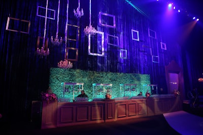 A wall of frames hung over a bar backed with greenery.