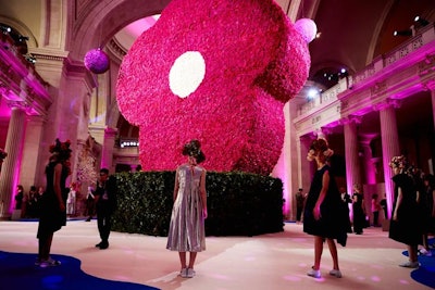 The central arrangement in the Great Hall, inspired by a Fall 2007 Comme des Garçons appliqué violet collection, took event producer Raúl Ávila and his team a month to build off-site. It was disassembled and brought into the Met the Sunday before the gala, where a large crew applied the flowers over an 18-hour period. The base of the desk was trimmed in boxwood with camellias.