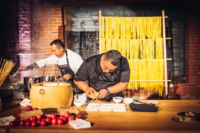 The dish is prepared at an action station, with fresh pasta displayed behind the chefs.