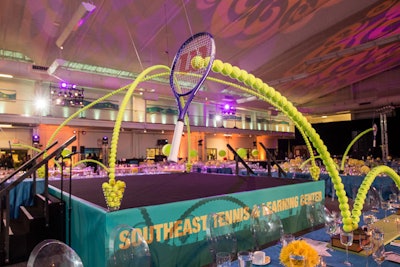 In November 2014, RJ Whyte Event Production produced the grand re-opening celebration, titled “Tennis Shoes, Ties, and After 5,” for the Southeast Tennis and Learning Center. The event was held in the 48,000-square-foot tennis center in Washington. Decor by Design Foundry included flying arches of tennis balls from hanging tennis racquets that bounced off of centerpieces of artfully stacked books. Large tennis balls were also stacked on glass vases to complement the high ceilings in the facility.