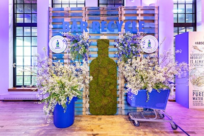 A step-and-repeat wall displayed a moss replica of the Absolut bottle against a wall of reclaimed wood, which featured the name of the event. White flowers poured out of a blue recycling bin and shopping cart.