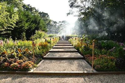 The Watermill Center used two distinct pathways for guest arrivals at its July 27 summer benefit. The main cocktails path took guests up a staggered walkway flanked by lush landscaping and hazy tiki torches into the forest of art installations; directly adjacent to the dinner check-in desk was a second path that took attendees through the main Watermill Center building (the former sole main entrance) and directly into the cocktail/silent auction space.