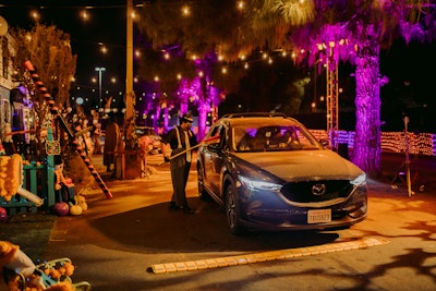 The drive-through also features 'door-to-door' trick-or-treating: Two streets are filled with themed house facades, and vehicles stop at each door where a costumed performer distributes wrapped candy from a safe distance using poles and buckets. Everyone is required to wear masks when car windows are down, and there is a staff of security guards and off-duty LAPD to enforce social distancing.